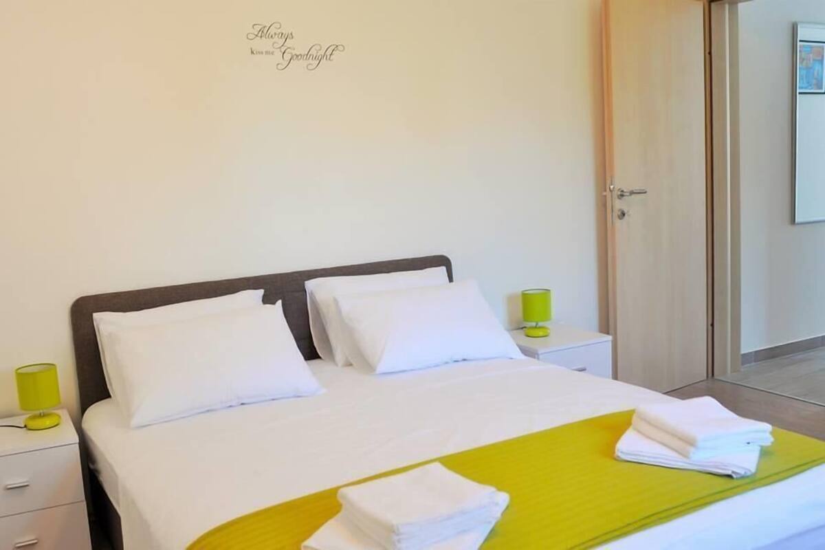 A neatly made bed with white linens, a yellow throw blanket, and a stack of white towels on a yellow bed runner.
