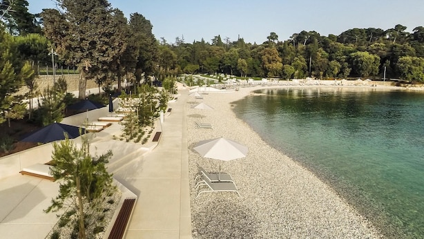 A pebble beach with umbrellas and lounge chairs, surrounded by trees and a clear view of the water. - Apartment Acimov A1 Two Bedrooms (Rovinj)