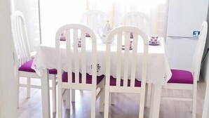 A dining area with white chairs and a table, a white refrigerator, and a window with sheer curtains. - Apartments Cagalj (Trogir)