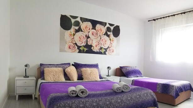 A bedroom with two beds, a nightstand with a lamp, a wall-mounted painting of roses, and a window with curtains. - Apartments Cagalj (Trogir)