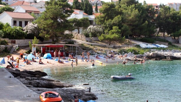 A beach with people sunbathing and swimming, a small boat, and a red structure. - Apartments Sanader (Okrug)