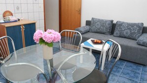 A dining area with a glass table, metal chairs, a vase of pink flowers, a small white table, and a grey sofa with patterned cushions. - Apartments Sanader (Okrug)