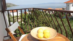 A balcony with a wooden chair and a wicker bowl containing lemons, overlooking a coastal view with houses and greenery. - Apartments Sanader (Okrug)