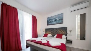 A hotel room with a large bed, red curtains, a painting of the sea, an air conditioning unit, and a mirror.