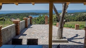 An outdoor patio with brick flooring, a table, and chairs, overlooking a scenic landscape with trees and a body of water.