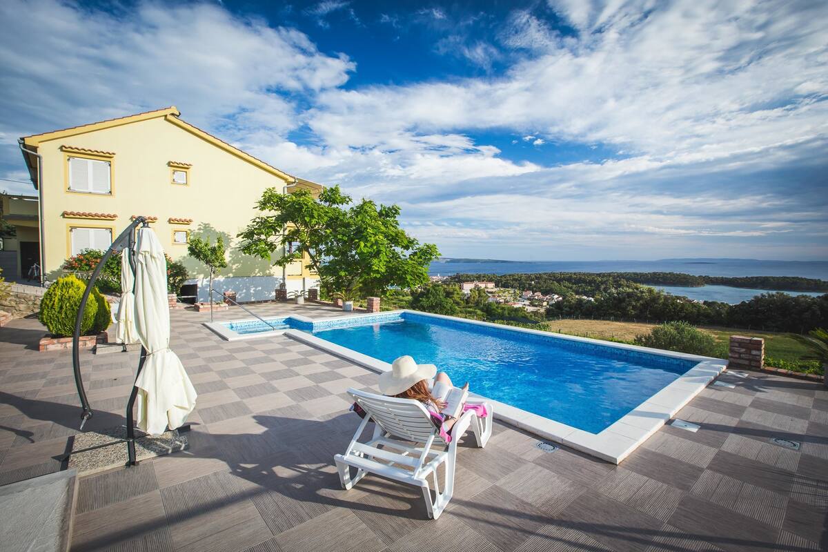 A poolside area with a view of the sea, a yellow house, and a person relaxing on a lounge chair.