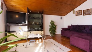 Un salon avec un plafond en bois, un téléviseur à écran plat, une vitrine contenant de la vaisselle, un canapé violet et une plante en pot.