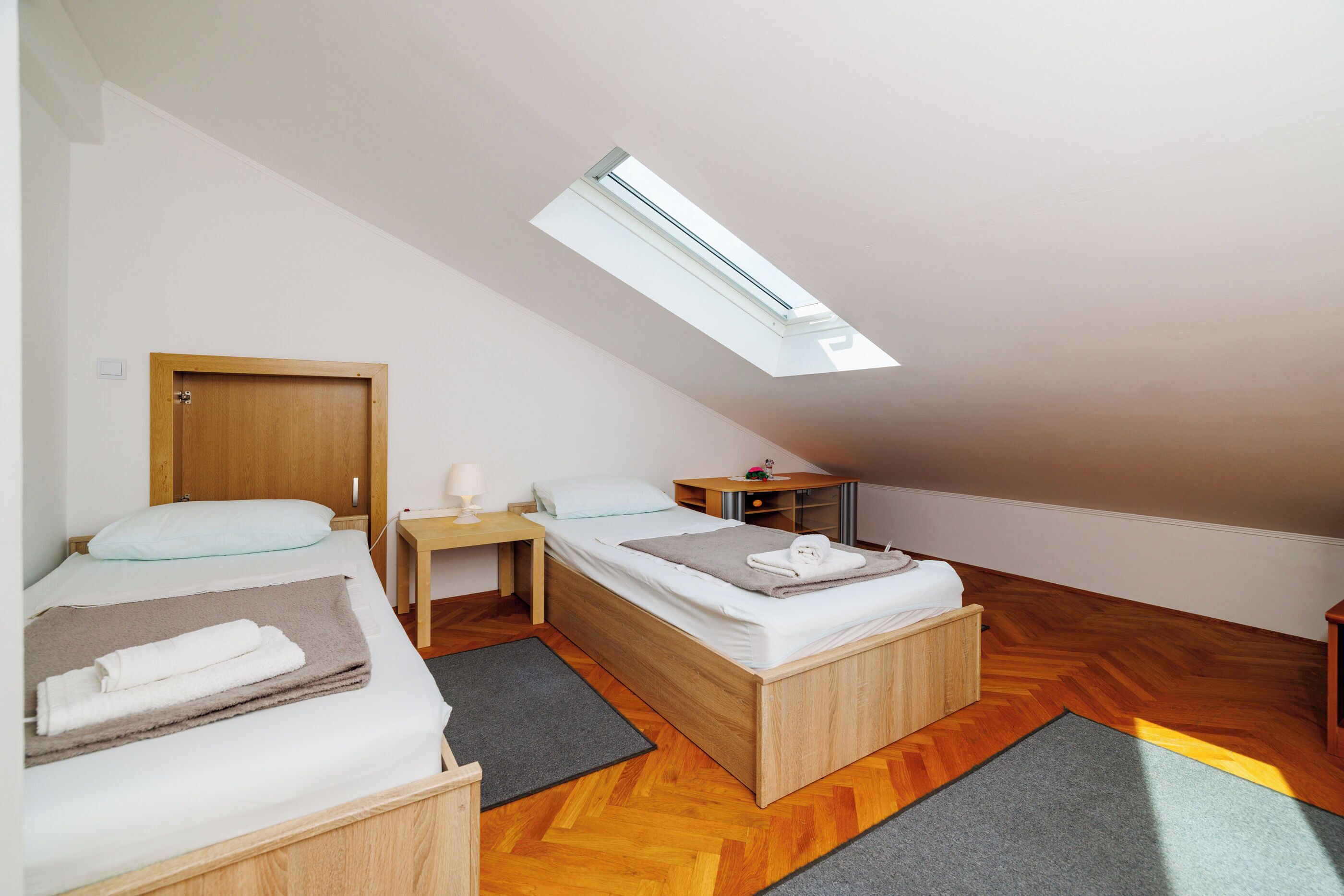 A small, modern room with a sloped ceiling, a skylight, two beds, a wooden wardrobe, a bedside table with a lamp, and a small desk.
