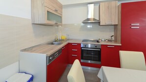 A compact kitchen with a microwave, oven, and sink. There are wooden cabinets and a red accent wall. A table and chairs are visible in the foreground. - Apartments Konta (Kastela)