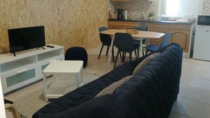 A living room with a black sofa, a TV, a small white table, and a black ottoman. There is a kitchen area with wooden cabinets, a refrigerator, and a window.