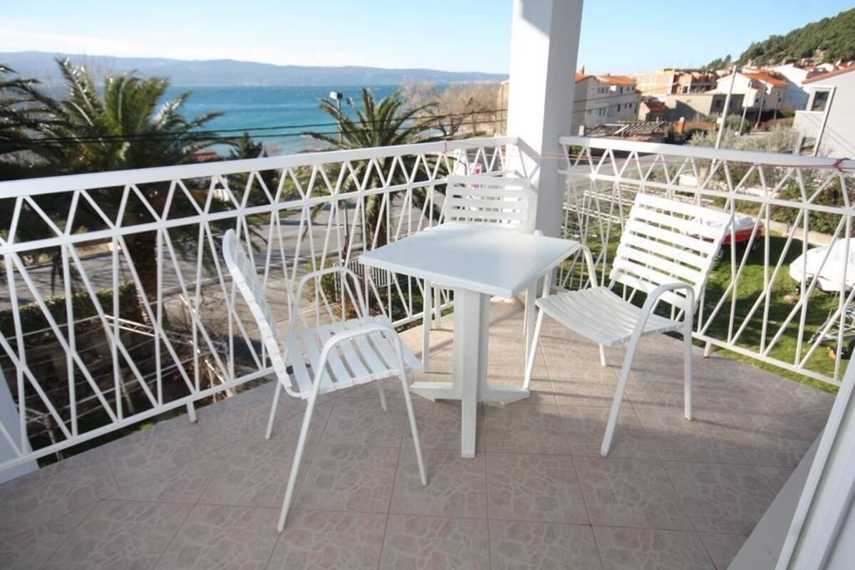 A balcony with a white table and chairs, a white railing, and a view of the sea and buildings.