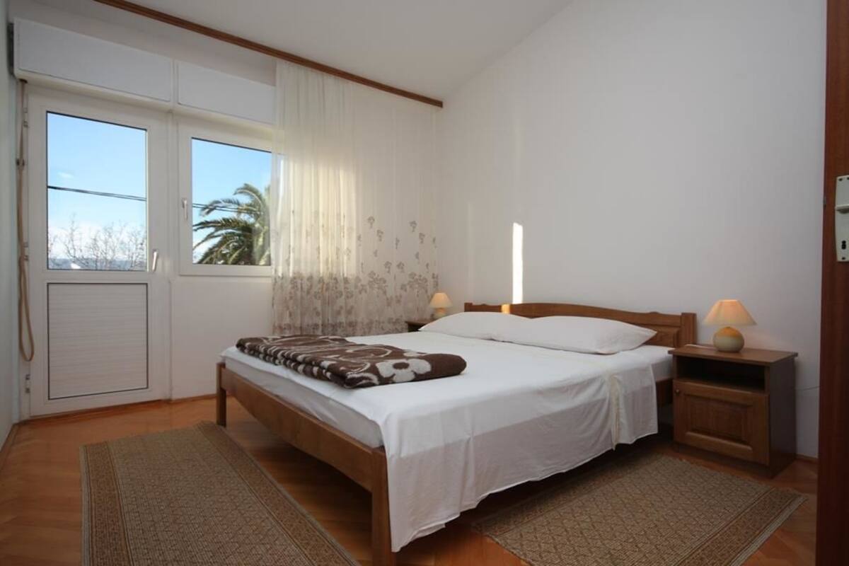A bedroom with a wooden bed, white linens, a nightstand with a lamp, and a window with curtains.