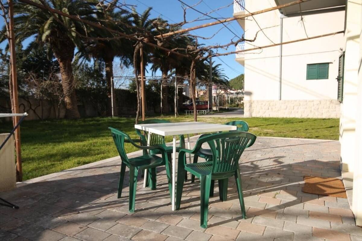 An outdoor patio with a table and chairs, surrounded by a garden with palm trees and a white building in the background.