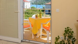 A room with a sliding glass door leading to a balcony with a yellow table and chairs, a potted plant, and a view of a body of water and hills. - Apartments Anton (Marina)