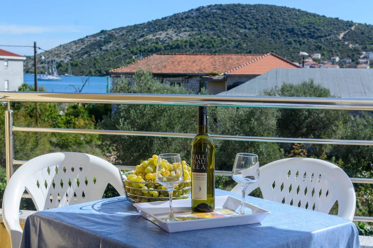A table with a bottle of wine, glasses, and a bowl of grapes on a balcony overlooking a scenic view of a hill and water.