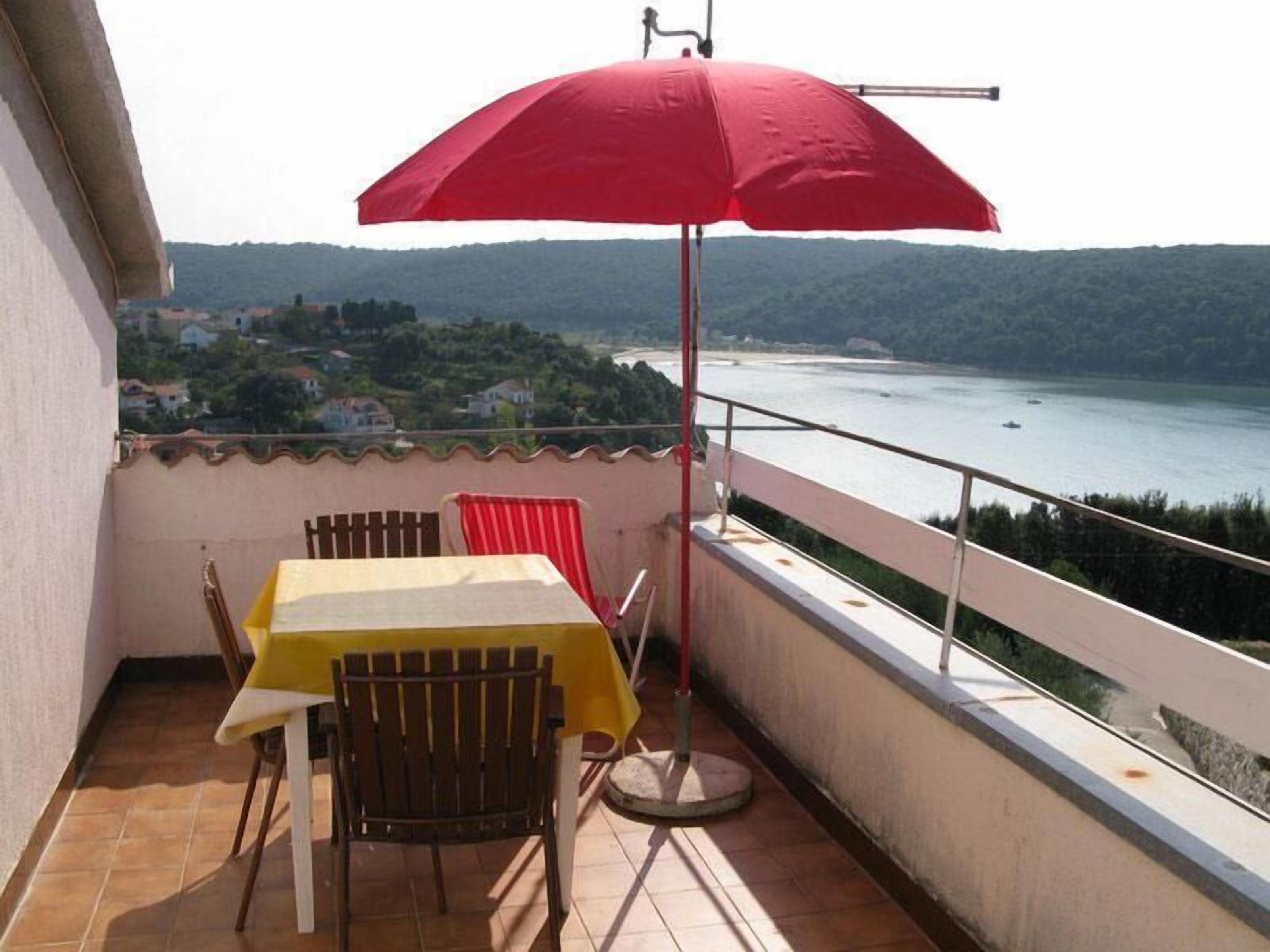 A balcony with a table and chairs, a red umbrella, and a view of a body of water and hills.