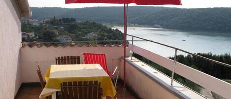 A balcony with a table and chairs, a red umbrella, and a view of a body of water and hills.