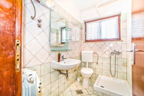 A bathroom with a white sink, toilet, and bathtub, a glass shelf above the sink, and a window with blinds. - Apartment Belušić / Three Bedrooms A1 (Fazana)