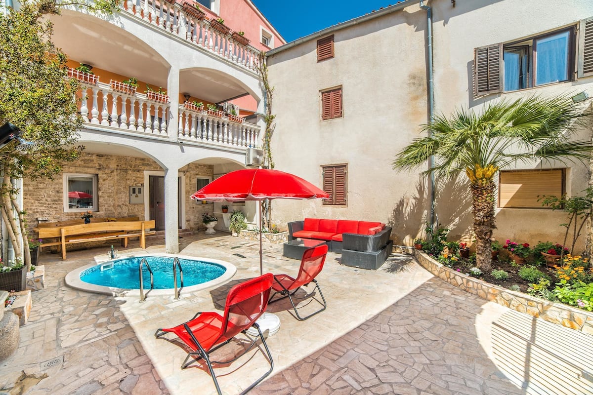 A courtyard with a swimming pool, red lounge chairs, a wooden bench, and a red umbrella.