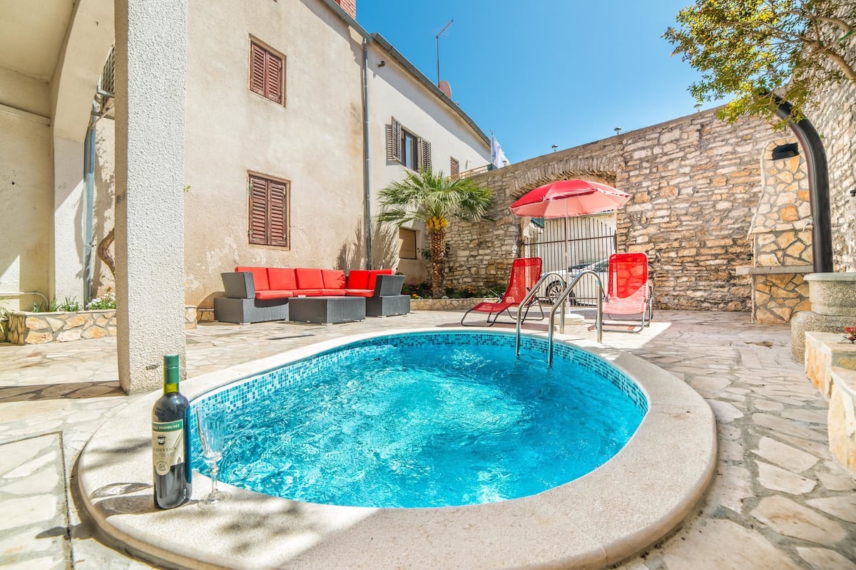 A swimming pool with a stone wall and a red umbrella, surrounded by outdoor furniture and a stone building.