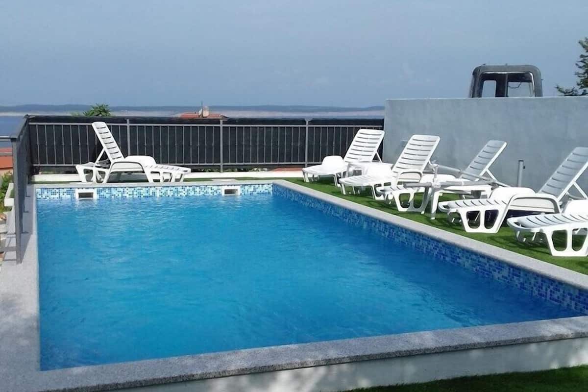 A rectangular swimming pool with a tiled edge, surrounded by white lounge chairs and a grassy area.