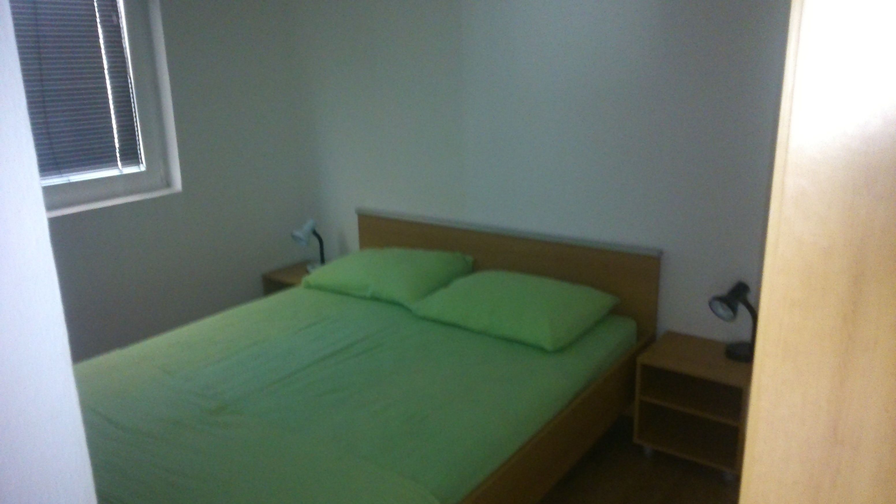 A small, single-bed room with a wooden headboard, a green bedspread, and a nightstand with a lamp.