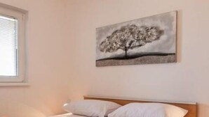 Una cama bien hecha con sábanas blancas y dos toallas enrolladas, una mesita de noche de madera y un cuadro de un árbol en la pared.