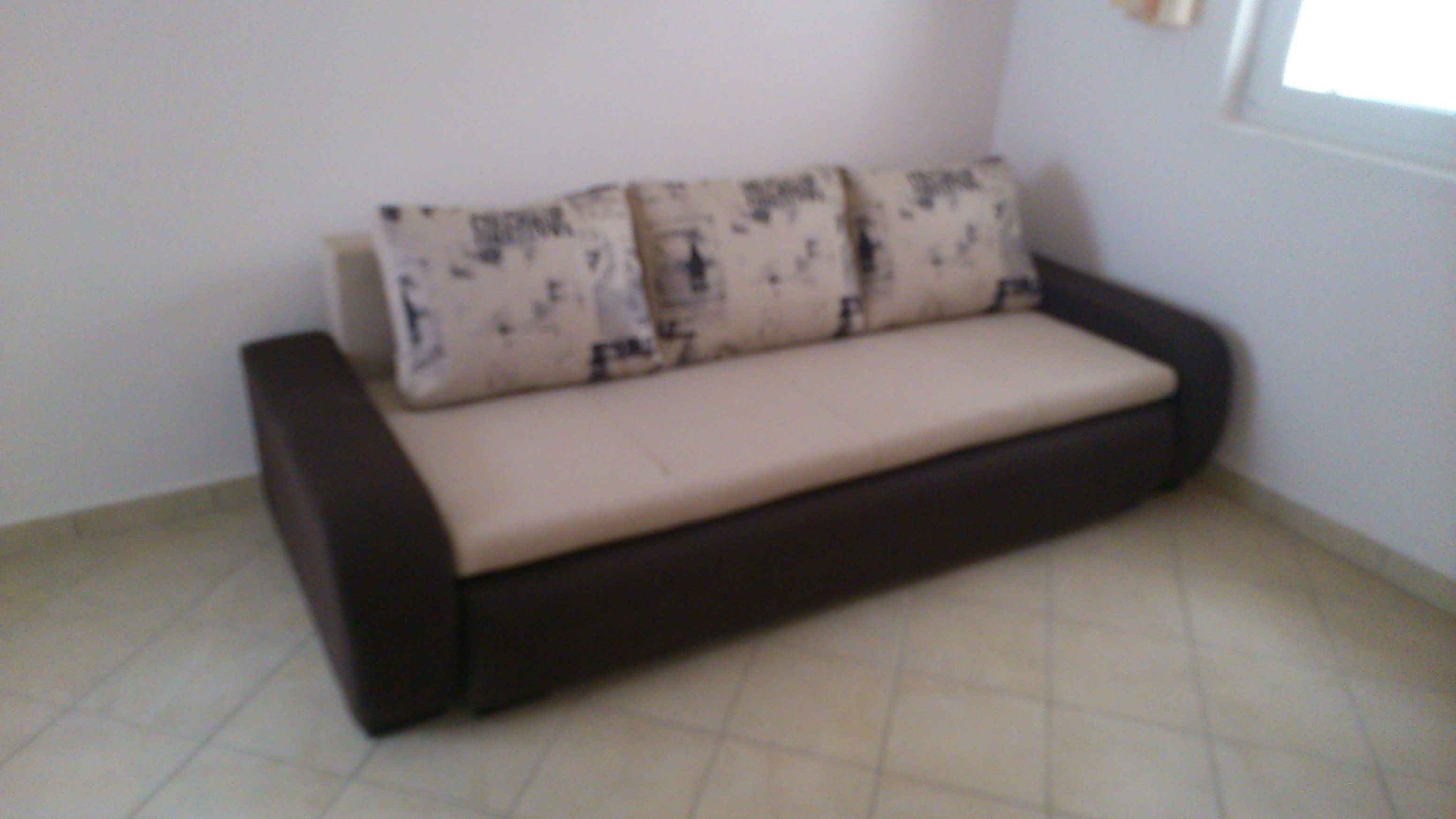 Un sofá pequeño de color oscuro con un cojín de color claro, frente a una pared lisa, en una habitación con piso de baldosas.