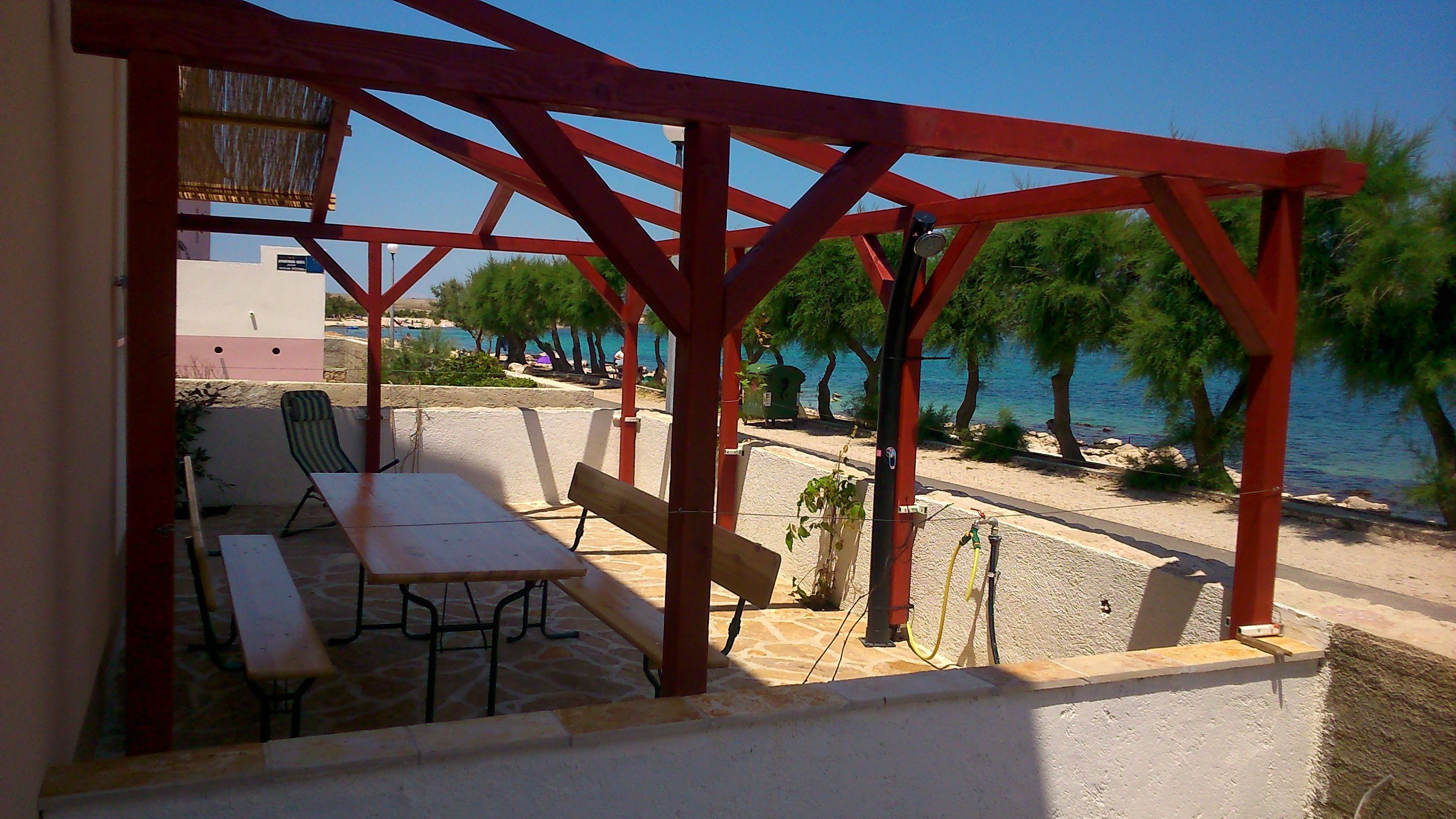 A covered outdoor seating area with a table and chairs, overlooking a beach with trees and a clear sky.