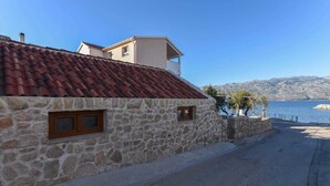 Un edificio de piedra con techo de tejas rojas, una ventana pequeña y vista a un cuerpo de agua y montañas al fondo.