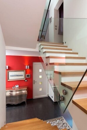 Eine moderne Treppe mit Glastrennstäben und Holzsitzgelegenheiten führt zu einem Raum mit einer roten Akzentwand und einer silbernen Kommode.