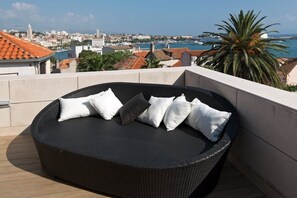 Eine schwarze Rattan-Couch mit weißen und grauen Kissen auf einer Dachterrasse mit Blick auf die Stadt und einer Palme.
