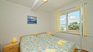 A bedroom with a bed, a nightstand, a lamp, a window with curtains, and a painting on the wall. - Apartments Vedrana (Dobrinj)
