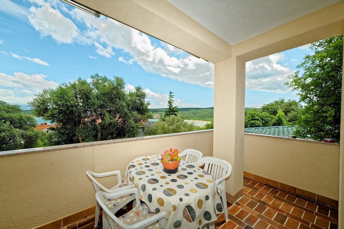 Un balcon avec une table et des chaises, une plante en pot et une vue sur la verdure et les bâtiments.