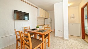 A compact kitchen with a dining area, including a TV mounted on the wall, wooden cabinets, and a tiled floor. - Apartments Vedrana (Dobrinj)