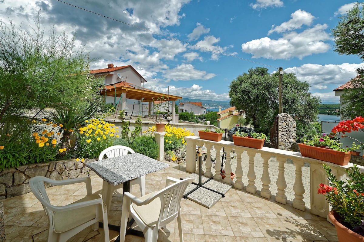 Une terrasse avec une table et des chaises, entourée de plantes et de fleurs en pot, d’où l’on peut admirer des maisons et une étendue d’eau au loin.
