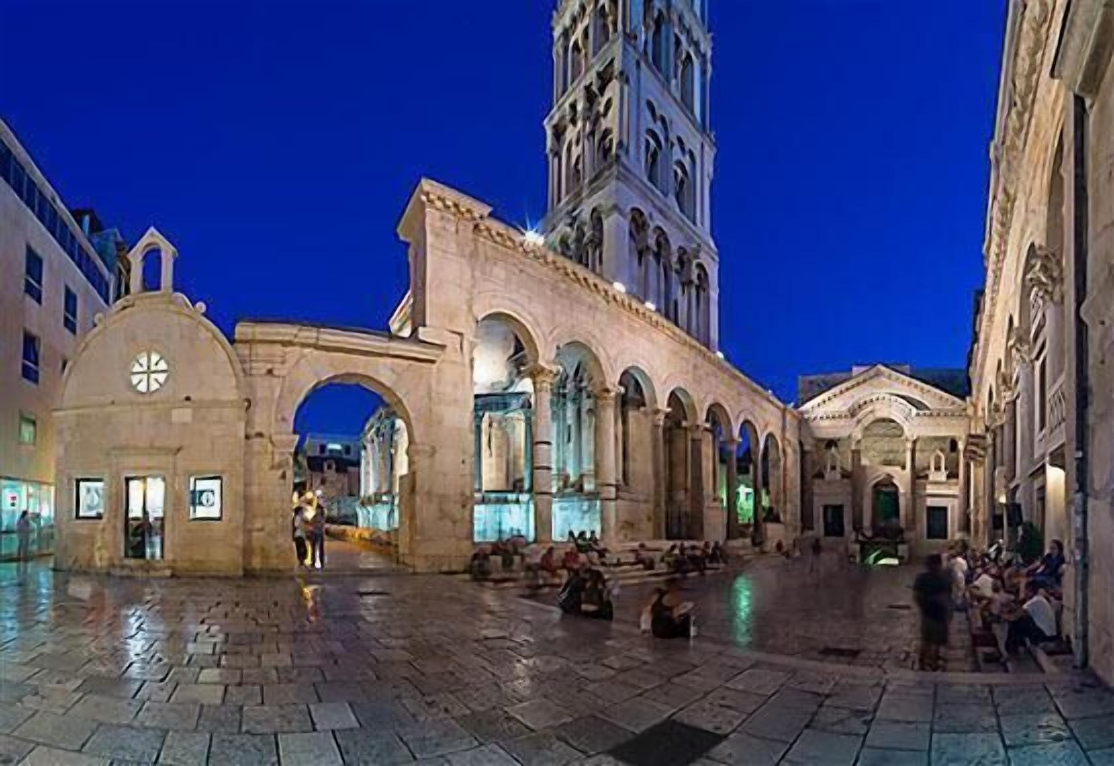 A historic square with a prominent clock tower and arches, surrounded by buildings with classical architecture.