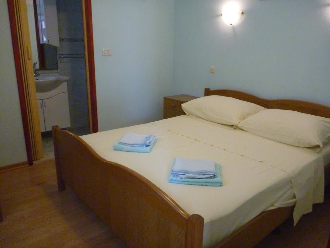 Un dormitorio con una cama de madera, una mesita de noche, una lámpara de pared y un baño visible a través de una puerta abierta.