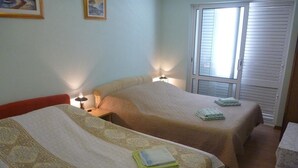 A hotel room with two beds, each with a patterned bedspread and pillows. There are bedside lamps, a window with blinds, and a small table with a lace tablecloth.