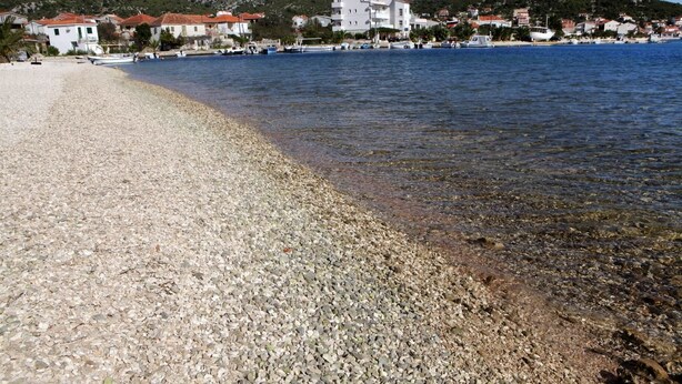 A pebbled beach with clear water, buildings in the background, and boats docked.