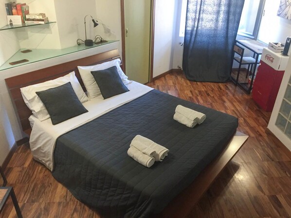 A neatly made bed with a dark quilt and two pillows, a glass shelf with books and a lamp, and a wooden floor.