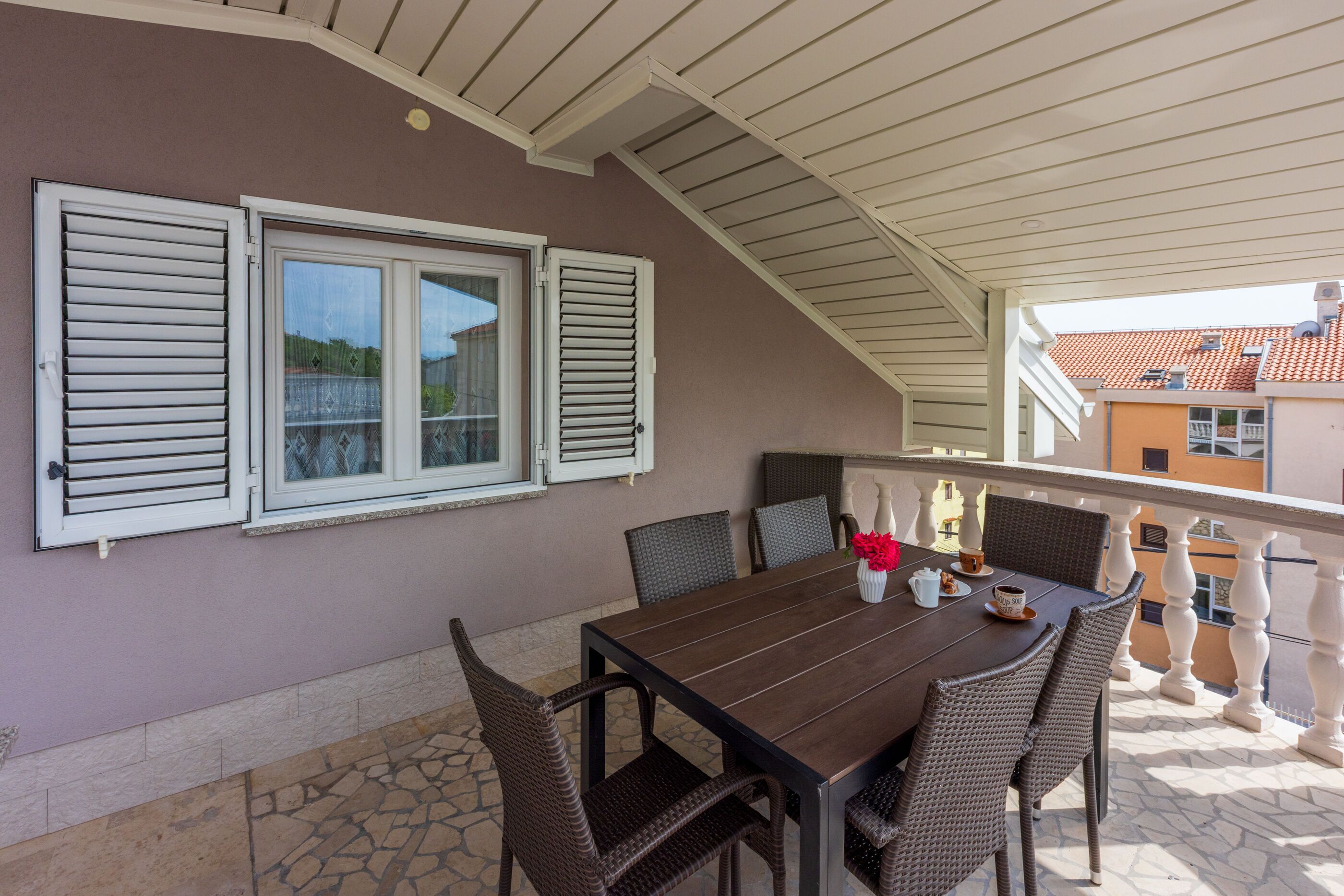 Eine Terrasse im Freien mit Holztisch, Korbstühlen, einem weißen Geländer und Blick auf angrenzende Gebäude.