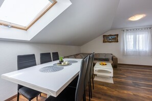 Un coin repas avec une table blanche, des chaises noires et une petite plante en pot. On aperçoit un canapé et une fenêtre avec des rideaux à l’arrière-plan.