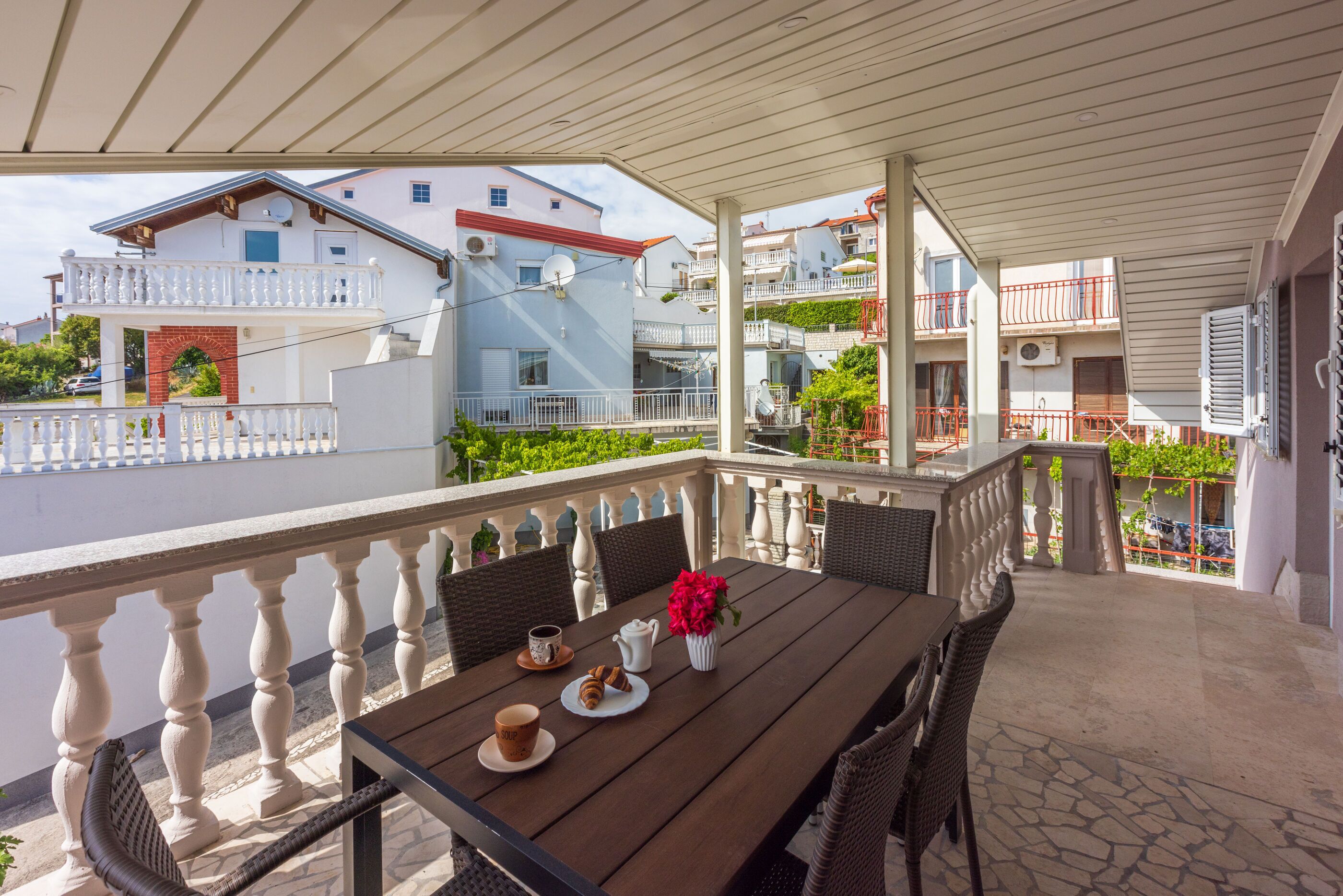A covered outdoor seating area with a table set for two, surrounded by wicker chairs, a white railing, and a view of neighboring buildings.