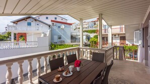 A covered outdoor seating area with a table set for two, surrounded by wicker chairs, a white railing, and a view of neighboring buildings. - Apartments Mirela (Crikvenica)