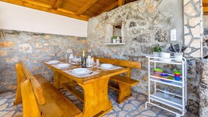 A wooden dining table set with plates, glasses, and a bottle, surrounded by benches, against a stone wall with a shelf above. - Holiday House Ivana (Malinska-Dubasnica)