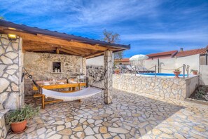 Une terrasse en pierre avec une table et des chaises en bois, un hamac et un espace piscine avec des chaises longues.