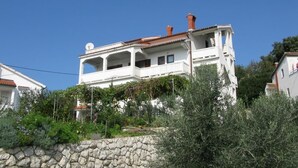 A two-story white building with a red-tiled roof, surrounded by greenery and a stone wall. - Apartments Mahić (Rab)