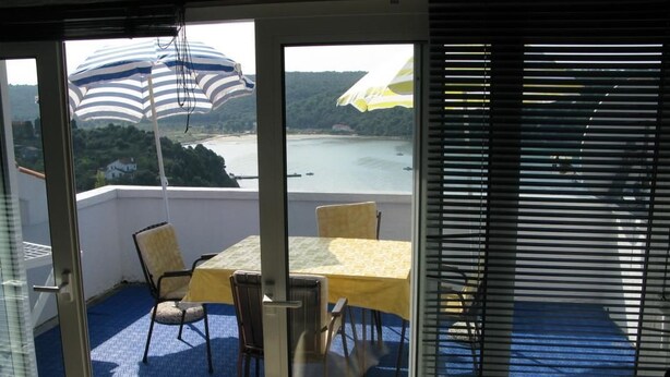 A balcony with a table and chairs, a view of a body of water, and a striped umbrella. - Apartments Mahić (Rab)