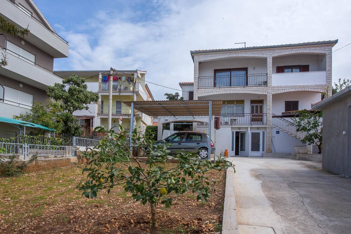 A multi-story residential building with balconies, a covered parking area, and a small garden with trees.